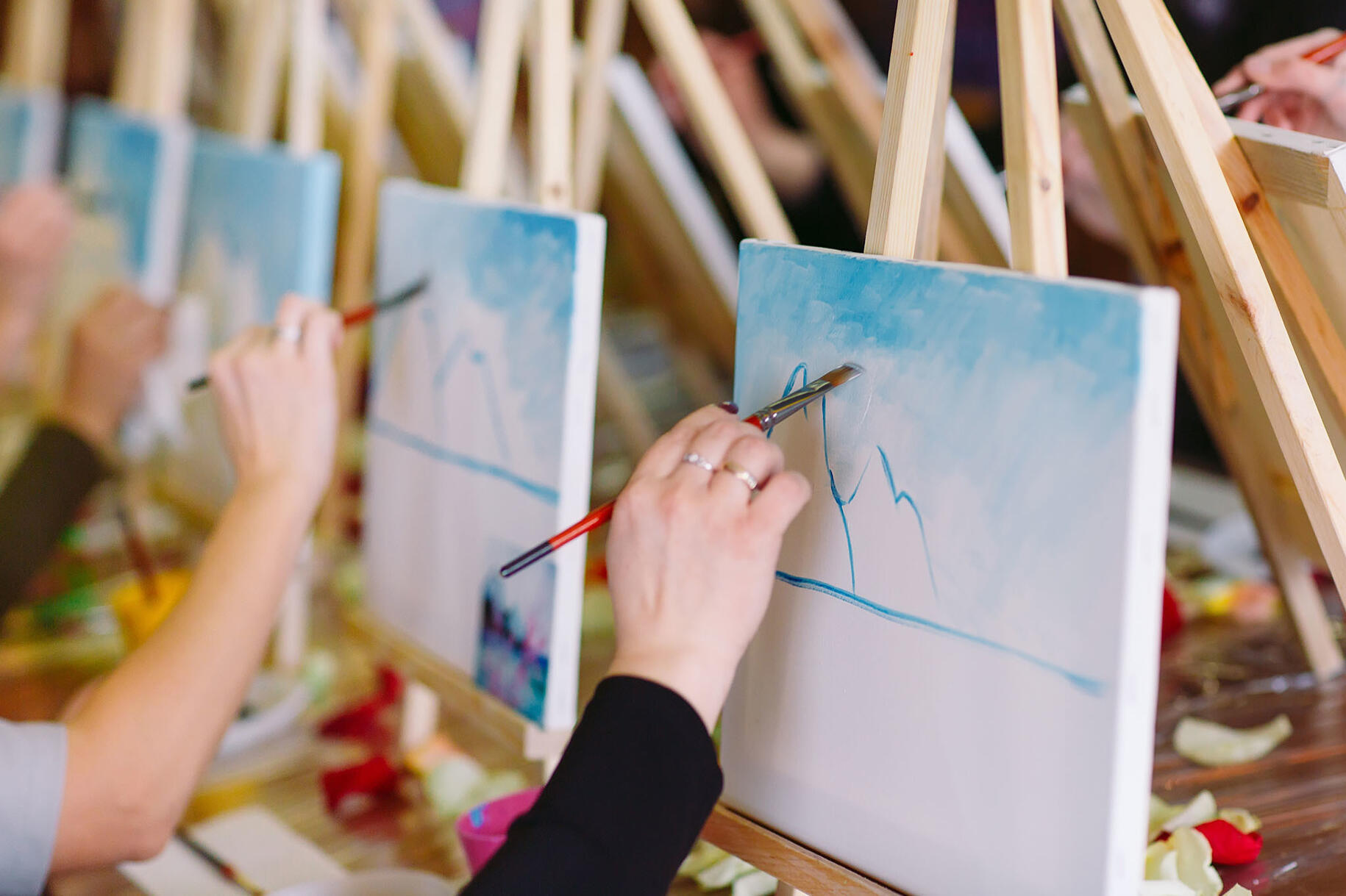Hands Painting Mountain Scene at Easel A hand painting on a canvas during an art session, with more art behind it.